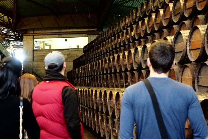 People standing in front of stacked wooden barrels in an industrial warehouse.