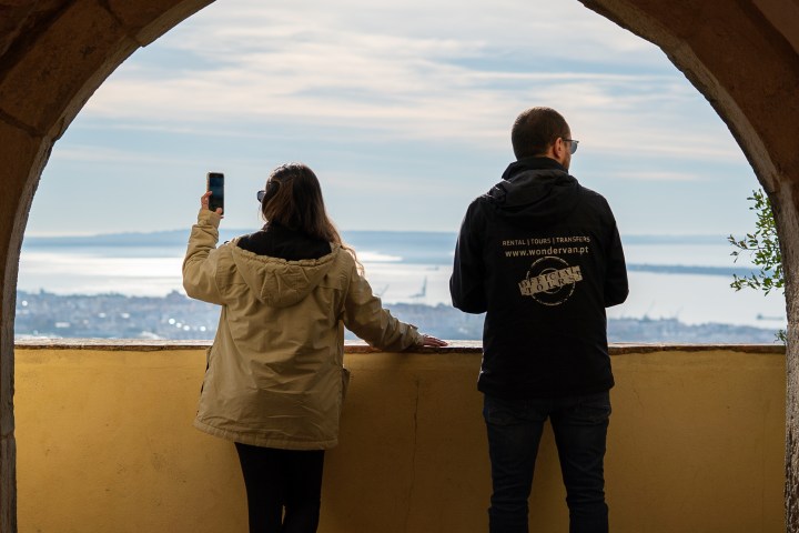 Two people standing under an archway, one taking a photo of a scenic sea view.