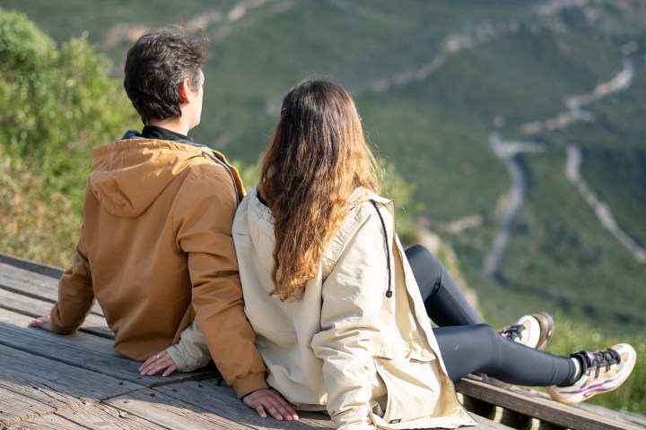 Two people in jackets sit on a bench overlooking a scenic landscape.