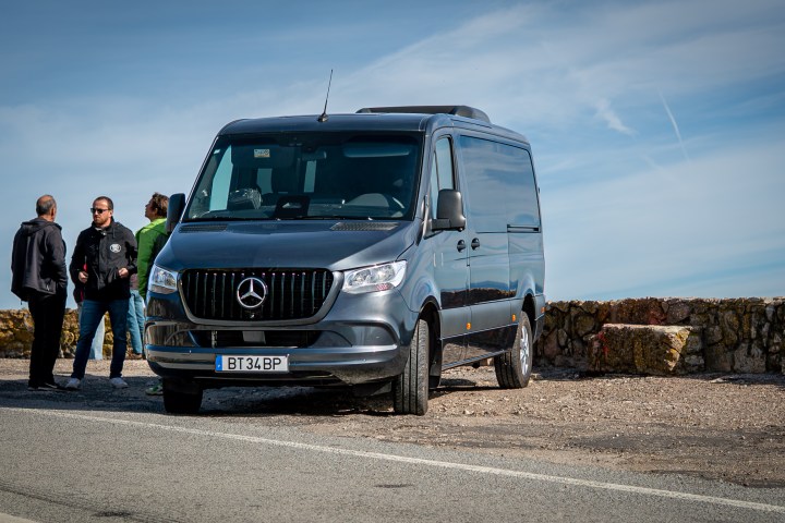 Gray van parked beside a road, with three people standing nearby.
