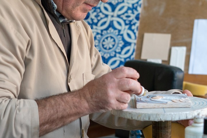Man in a beige coat painting a ceramic tile on a workbench.