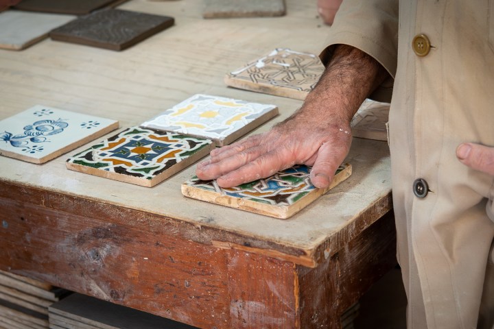 Person in beige coat touching decorative tiles on a wooden table.