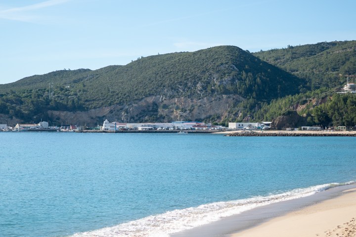 Beach with gentle waves, distant mountains, and buildings along the shore under a clear sky.