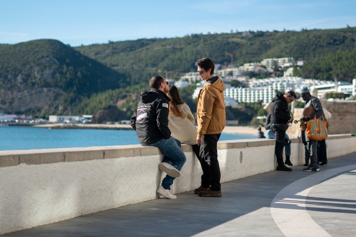 People socializing on a seaside walkway with mountains in the background.