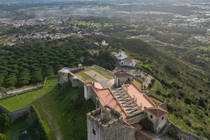 Aerial view of a historic castle with a tower, surrounded by greenery and overlooking a distant city and coast.
