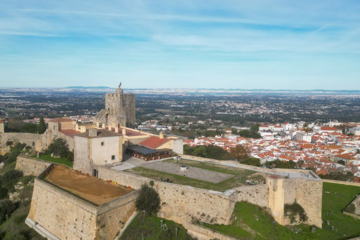 Aerial view of a large, historic stone fortress on a hill with cityscape in the background under a blue sky.