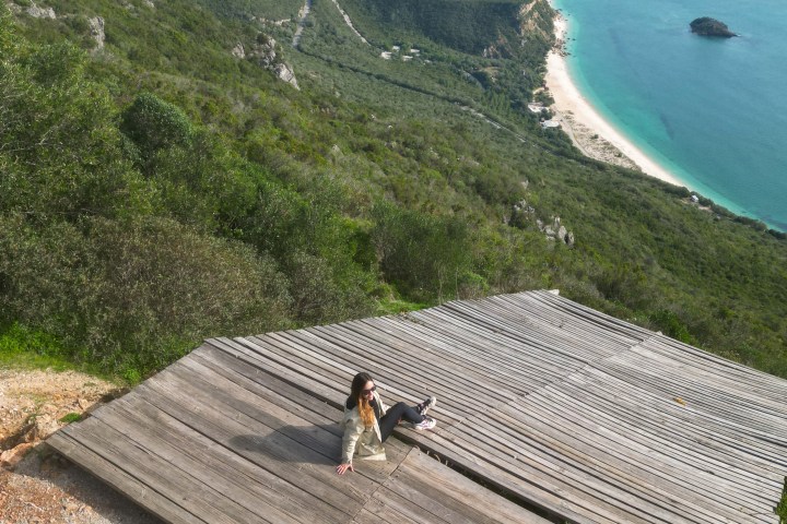 Two people take photos of a woman sitting on a wooden platform overlooking a coastal landscape.