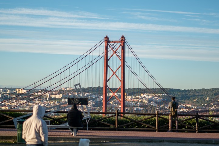 People view a large red suspension bridge overlooking a cityscape under a clear blue sky.