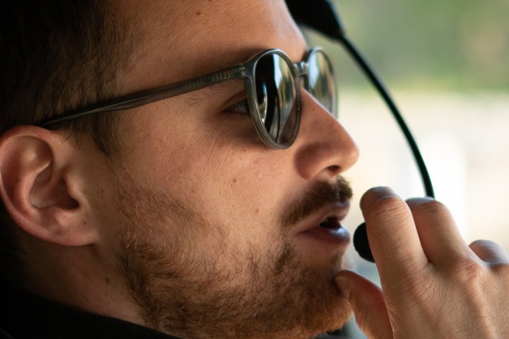 Man wearing sunglasses and headset, speaking into microphone in a vehicle.