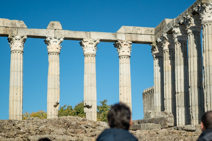Ancient stone columns with ornate capitals under a clear blue sky, viewed by three people.
