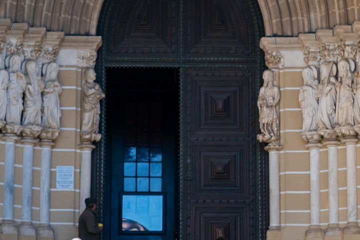 Two people standing on steps outside a large arched door with statues on either side.
