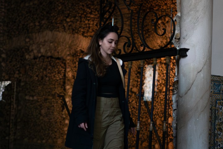 Woman in a dark coat walks down steps in a dimly lit ornate hallway.
