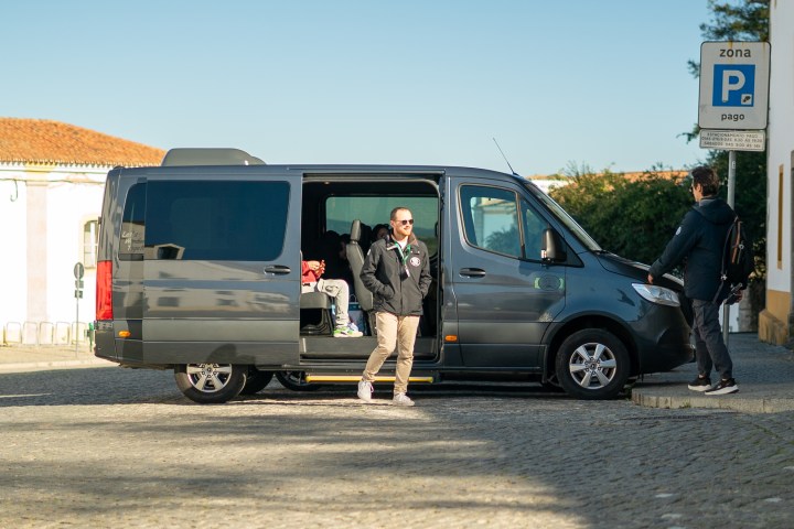 Man exits van parked in a cobblestone area; another person stands nearby.