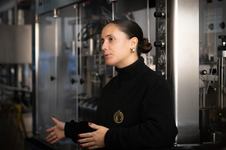 Woman in black jacket speaking in front of industrial equipment inside a factory.