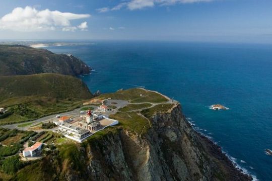 Aerial view of a lighthouse on a cliff overlooking the ocean under a clear blue sky.