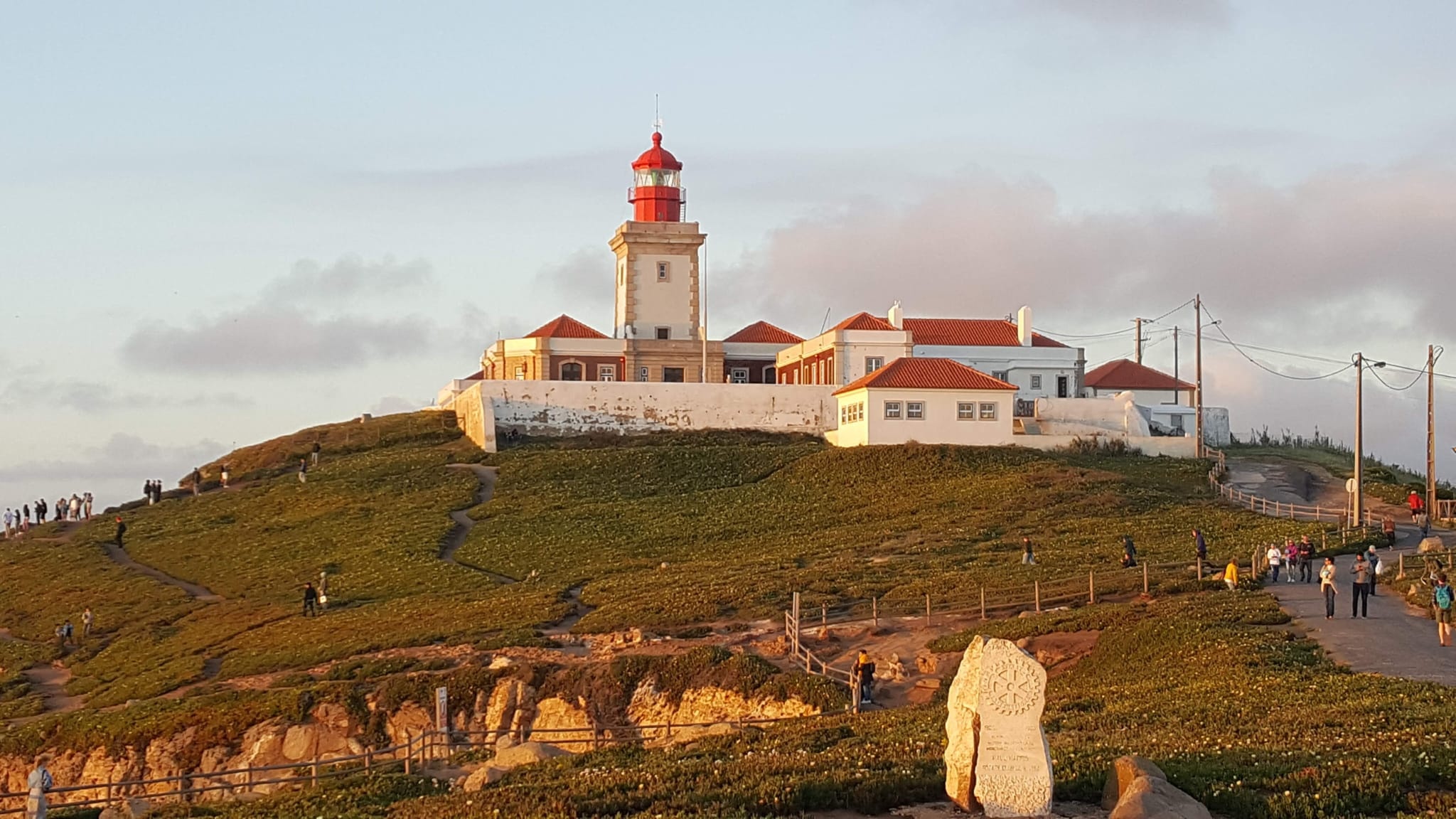 Lighthouse on a grassy hill at sunset with people walking around.