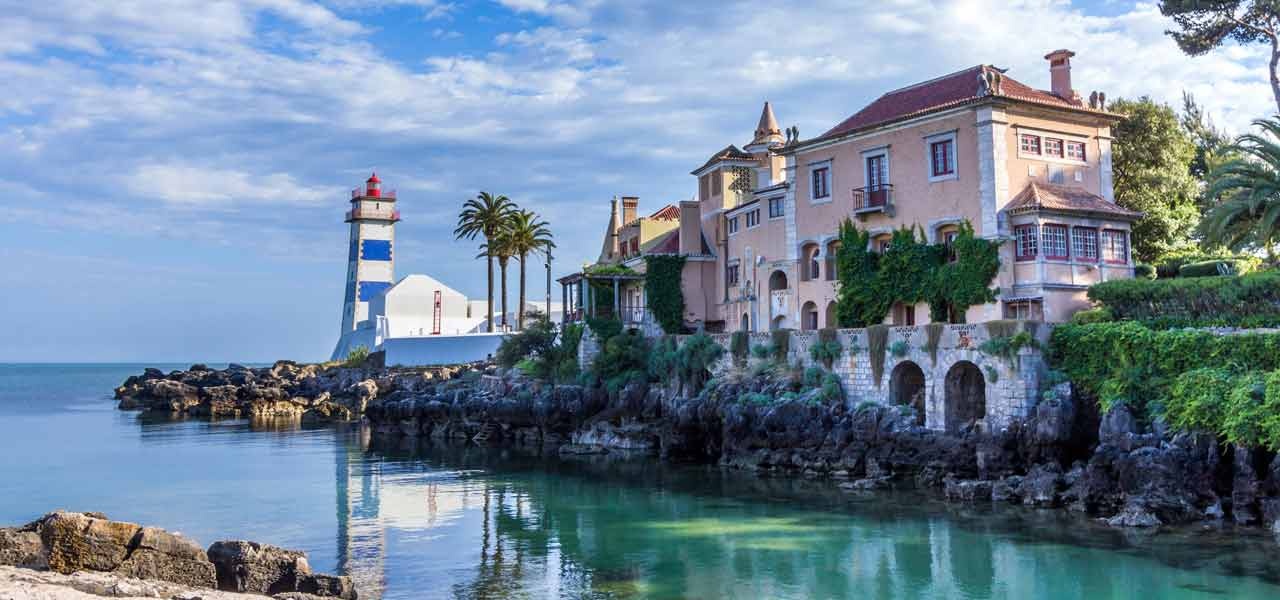 Coastal scene with lighthouse, historic building, palm trees, and rocky shoreline under a partly cloudy sky.