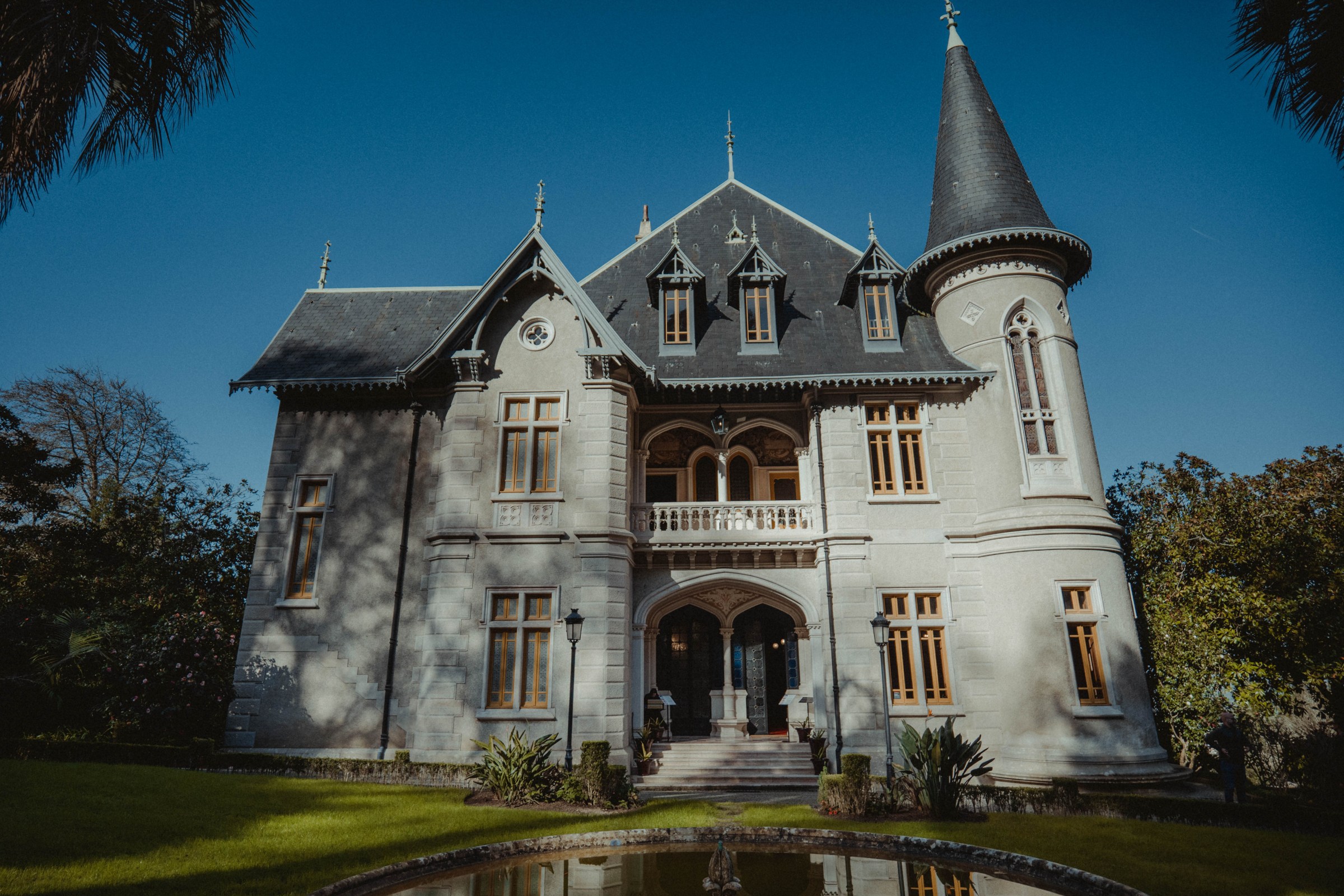 Victorian-style mansion with turret and steep roof, surrounded by greenery and a small pond.