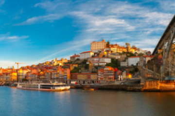 Panoramic view of a city by a river with a large metal bridge and colorful buildings under a blue sky.