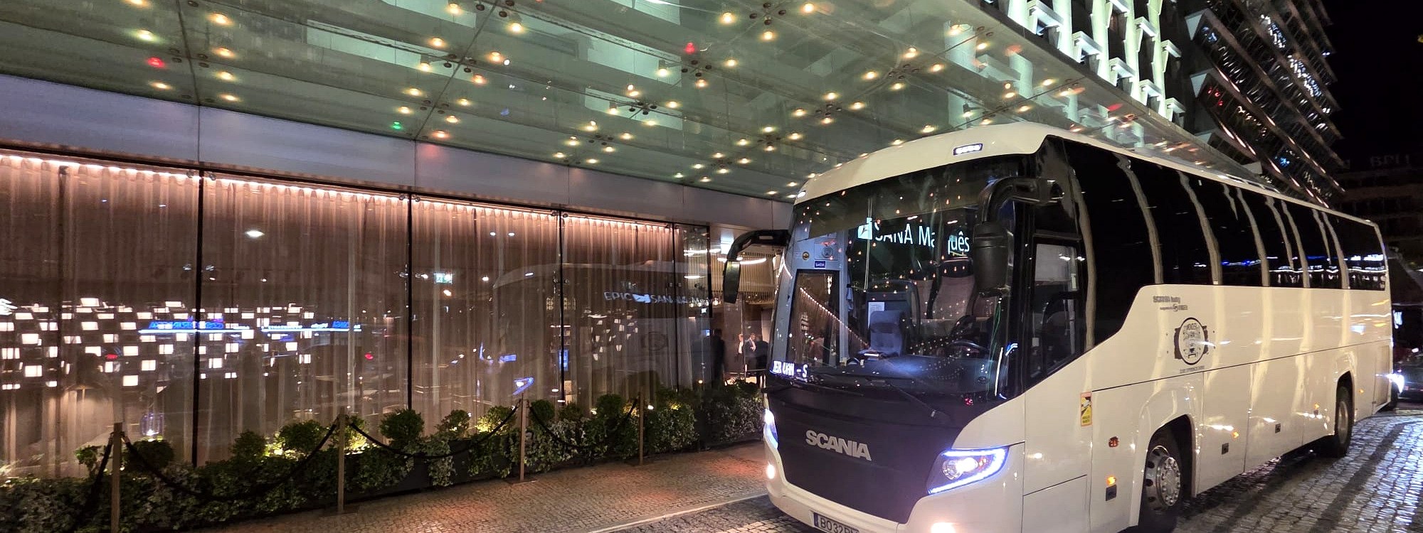 White tour bus parked outside a modern, glass-fronted building at night.